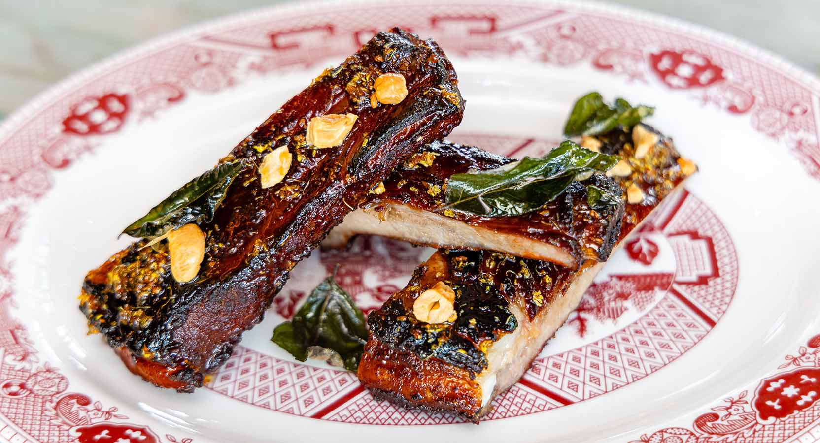 Glazed and grilled pork ribs topped with fried garlic slices and herbs on a red-patterned plate.
