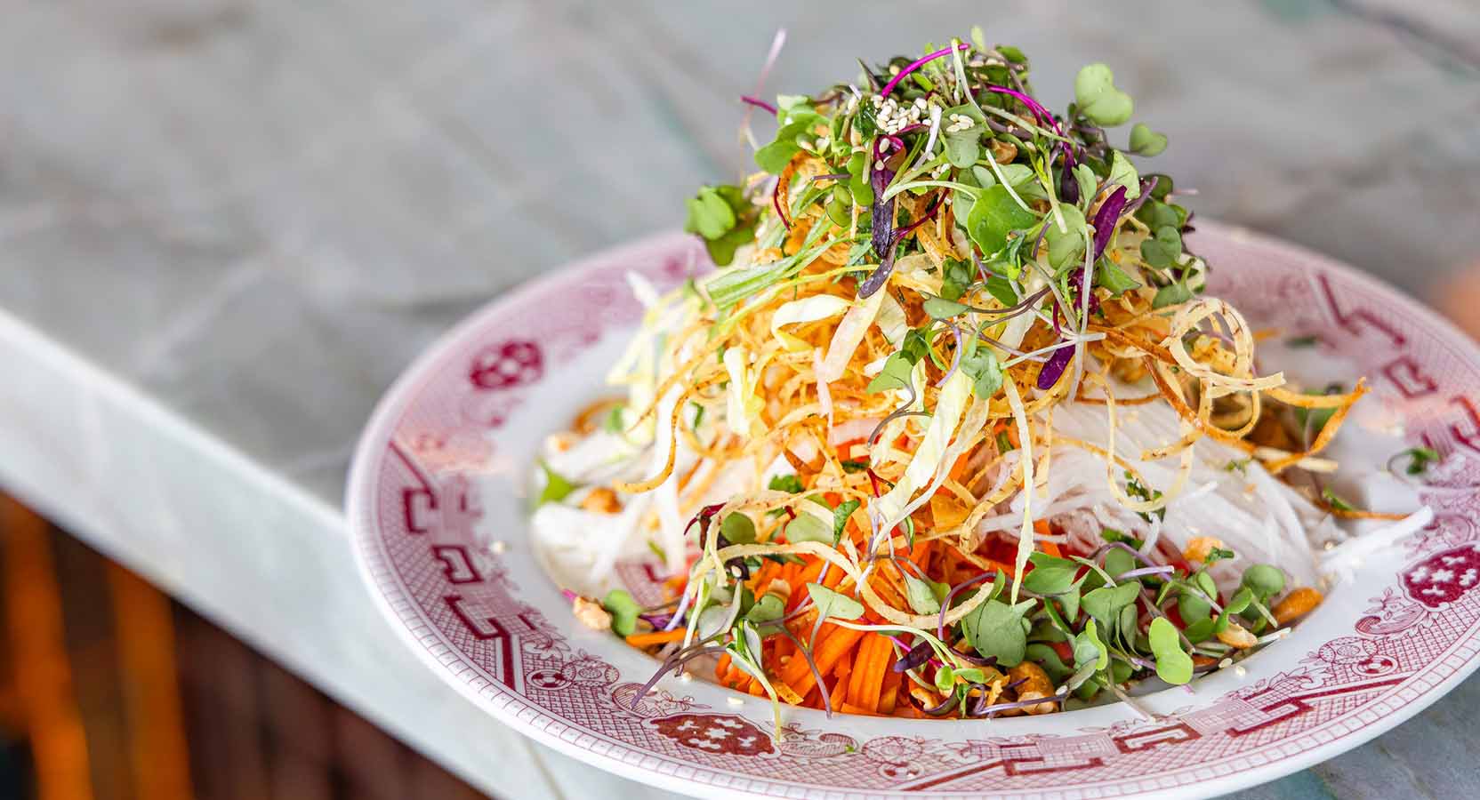 Mixed vegetable salad with shredded carrots, sprouts, microgreens, and crispy noodles on a porcelain plate.