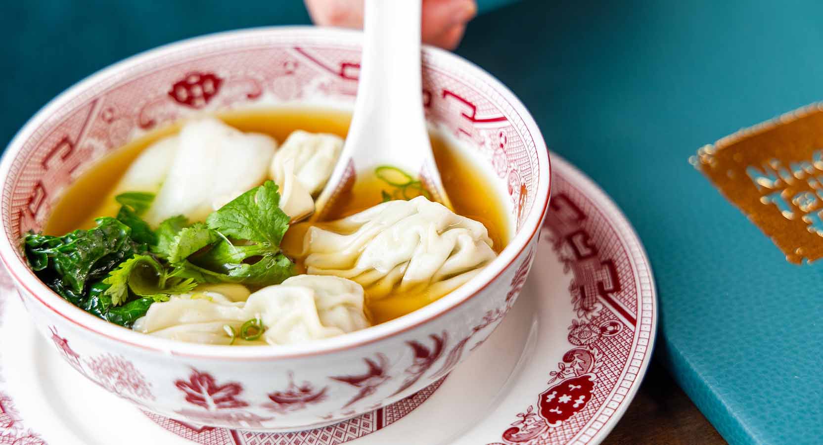 Clear broth with pork wontons, leafy greens, and cilantro served in a traditional red-and-white bowl.
