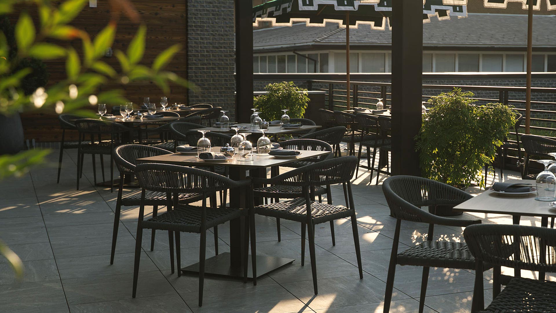 Outdoor patio seating with woven black chairs and square tables, surrounded by greenery and shaded by geometric umbrellas.