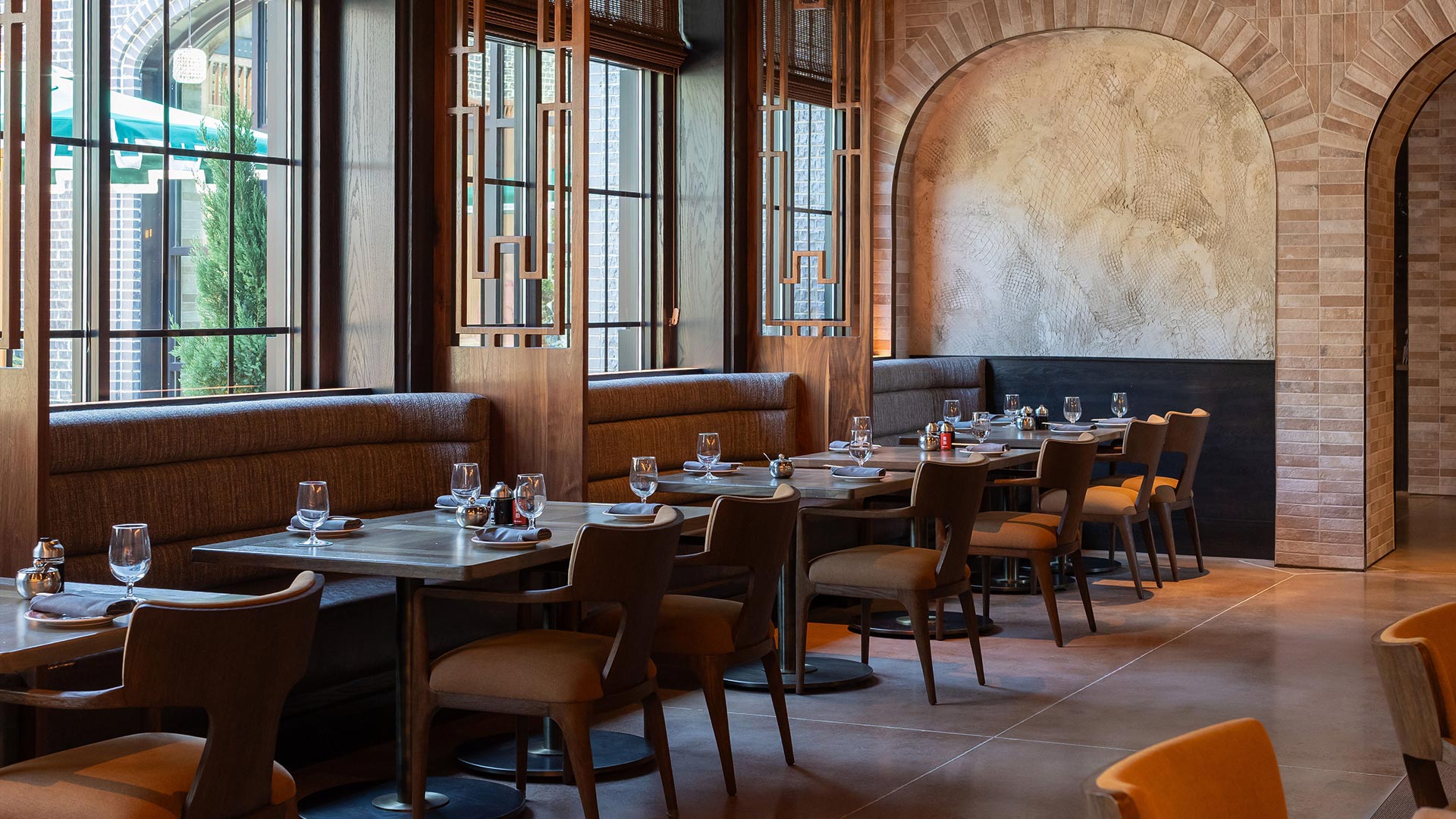 Sunlit dining space with arched stone wall, booth seating, and floor-to-ceiling windows framed by intricate wood dividers.