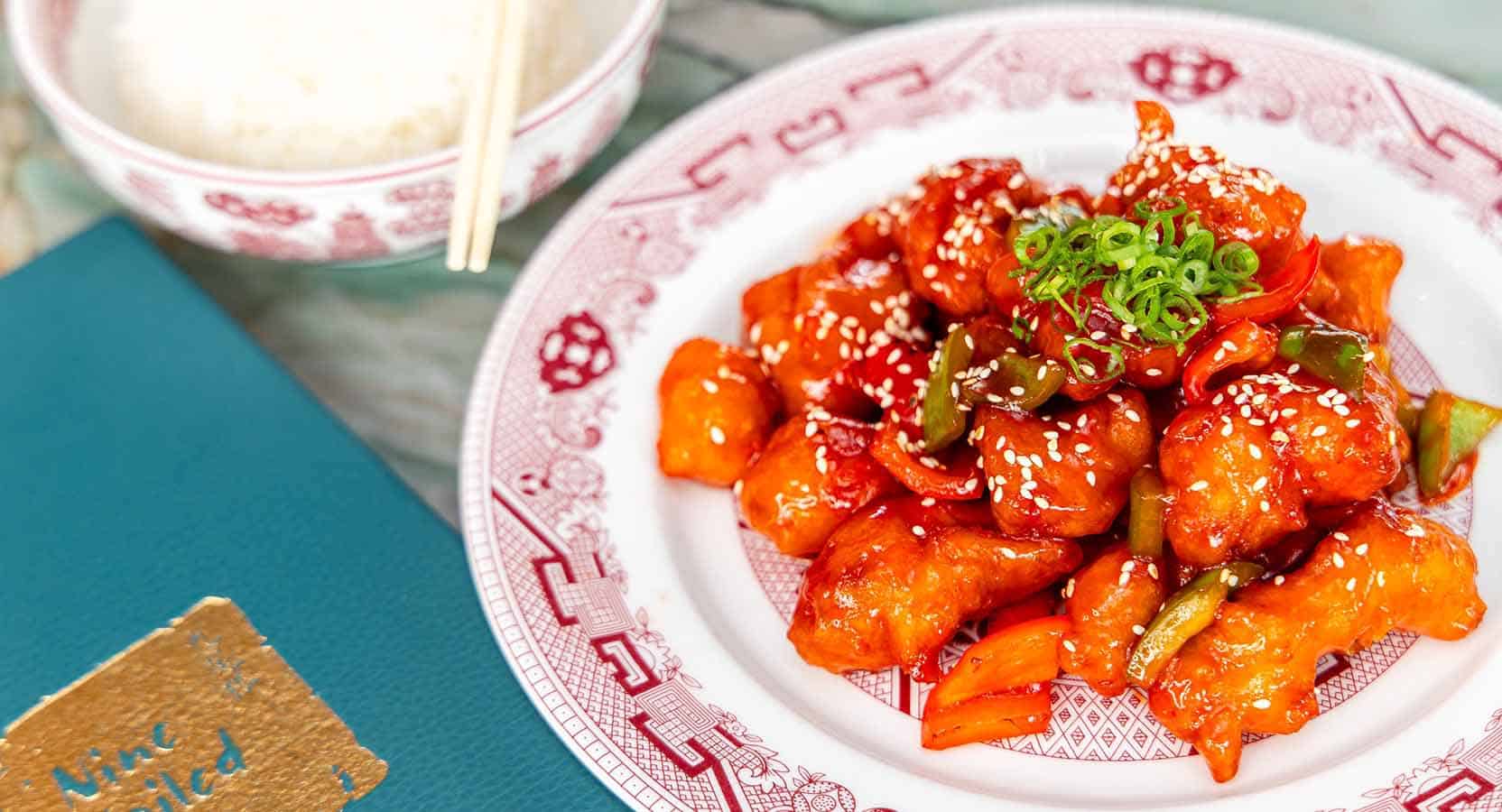 Spicy Chinese orange chicken dish on a decorative white plate with sesame seeds and chopped green onions, served with traditional Asian-style rice bowl, showcasing Asian cuisine and flavor.