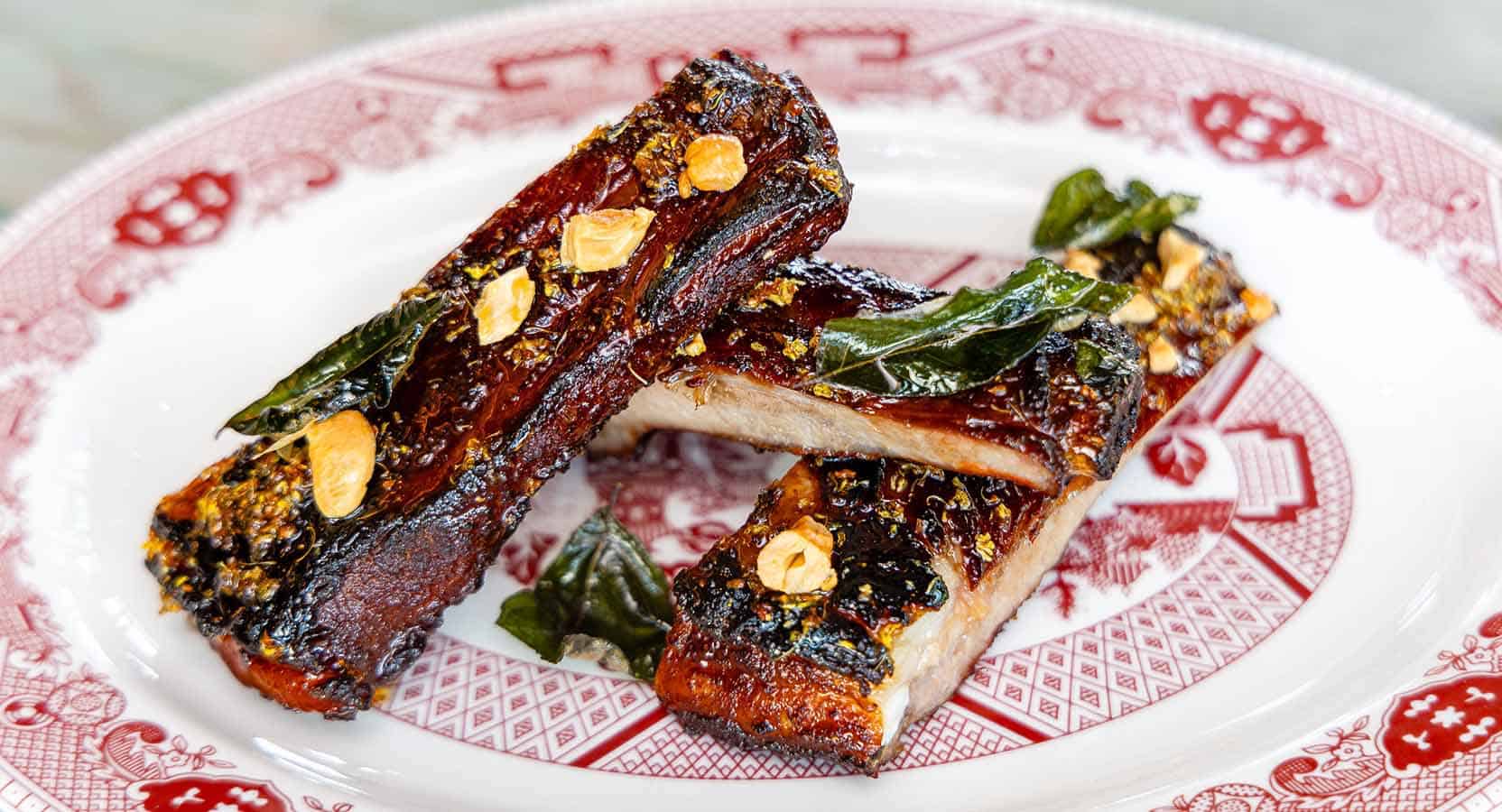Succulent glazed pork ribs with a crispy exterior, garnished with garlic, herbs, and roasted chili peppers, served on a traditional patterned plate for a flavorful Asian-inspired dish.